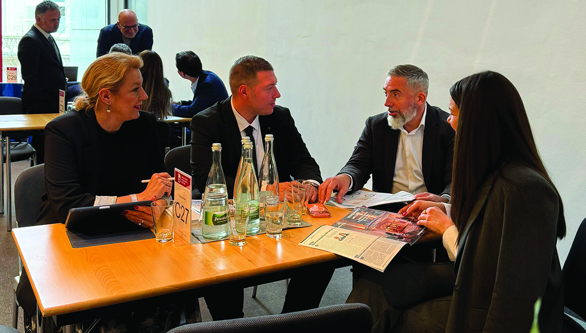 Grupa formalno obučenih ljudi razgovara sedeći za stolom na kojem su flaše vode, čaše i magazini